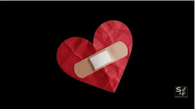 A crumpled red paper heart with a beige band-aid placed over it, set against a dark background. This visual metaphor represents a broken heart being superficially patched up, central to Katy Perry's "Bandaids" song.